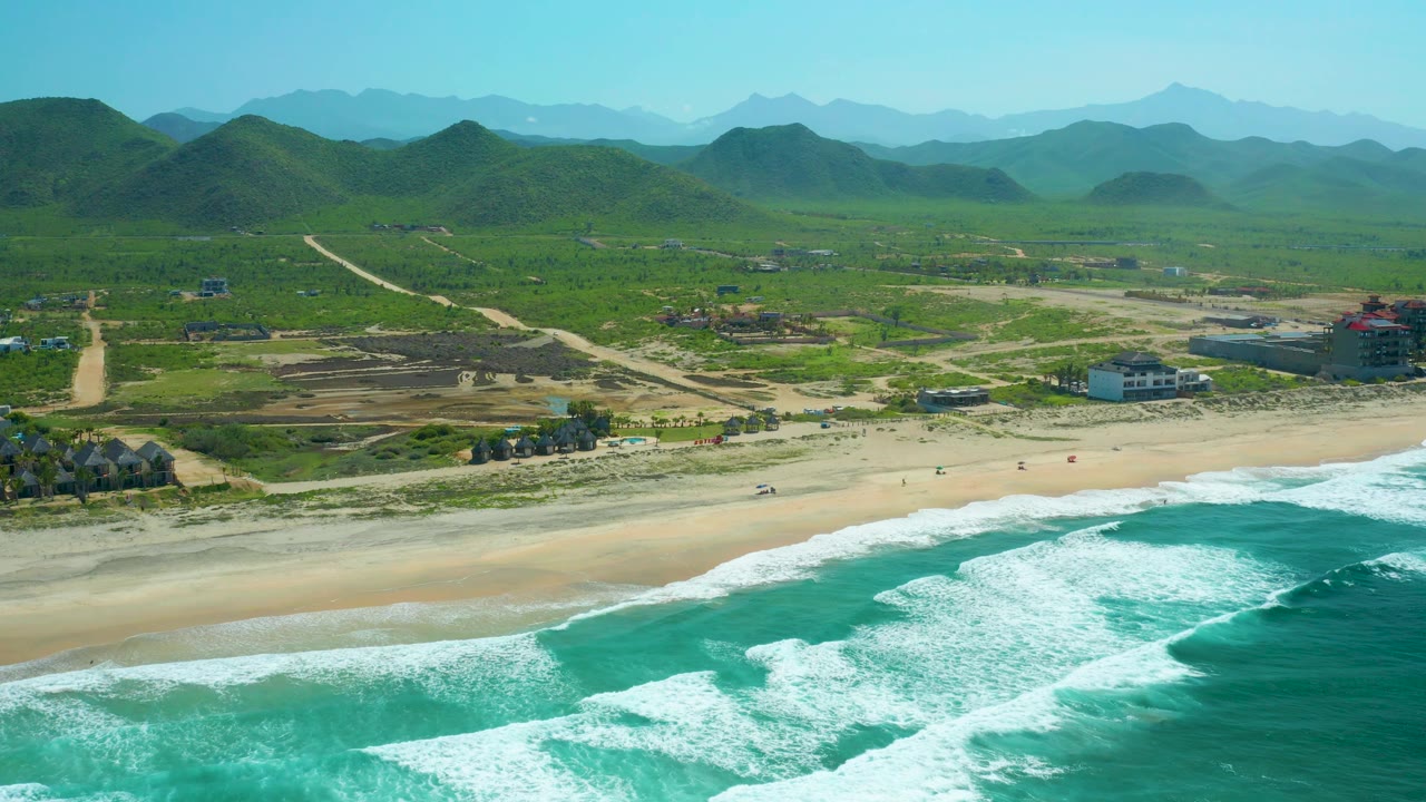 Aerial frame over Cerritos Beach showing a broad white wave band, sandy shoreline, beachfront structures, construction changes inland, green hills, and no visible people.