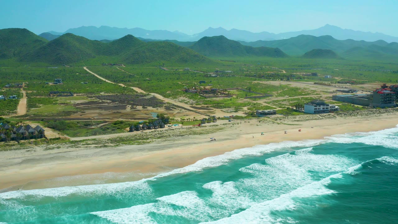 Cerritos Beach view with surf washing toward the lower frame, empty sand, low structures near the beach, exposed construction ground, green hills, and no visible people.