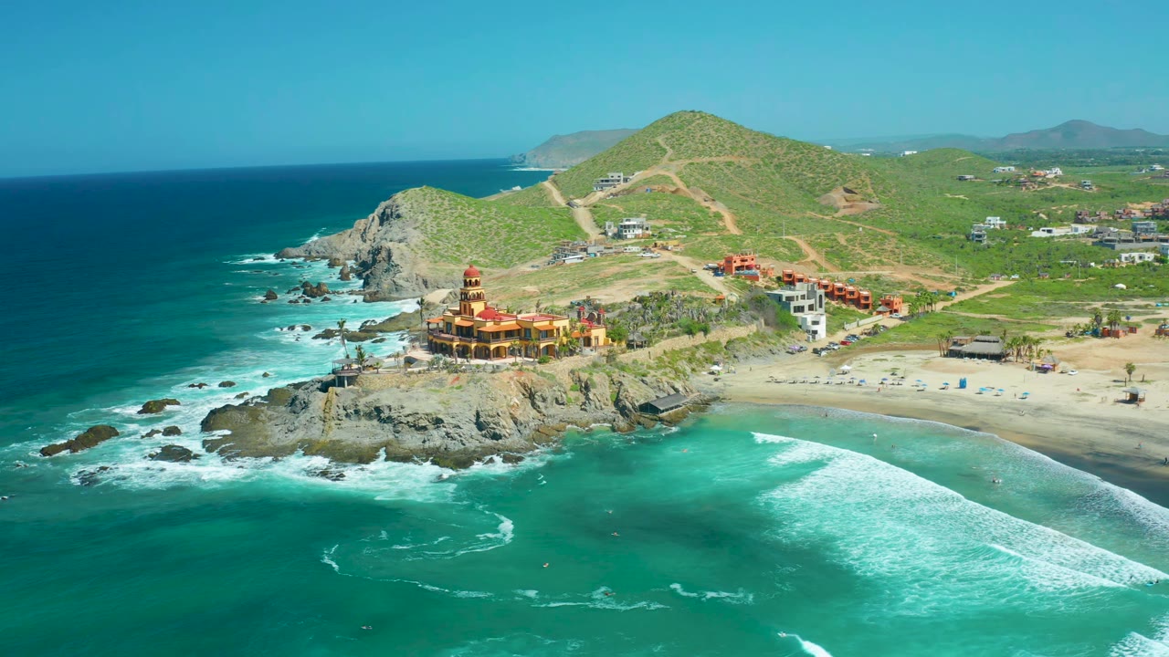 Wide aerial coastline with surf, rocky headland, beach, and hillside roads.