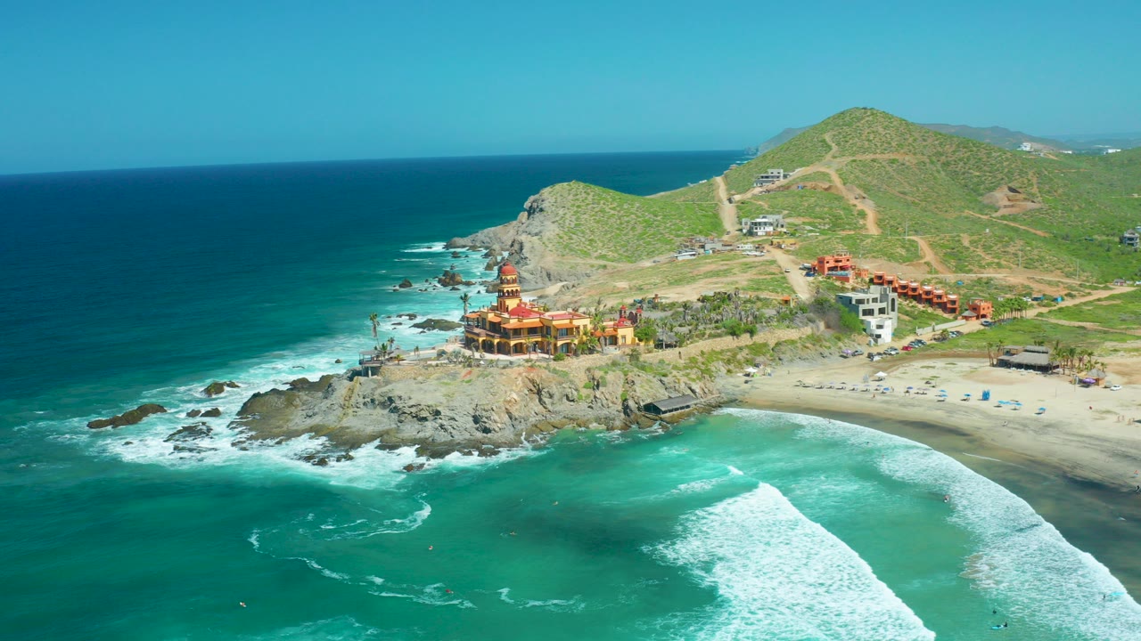 Aerial frame over Cerritos Beach, Baja California Sur, with turquoise water in front, a rocky point and temple-like building near center, green hills behind, and no visible people.