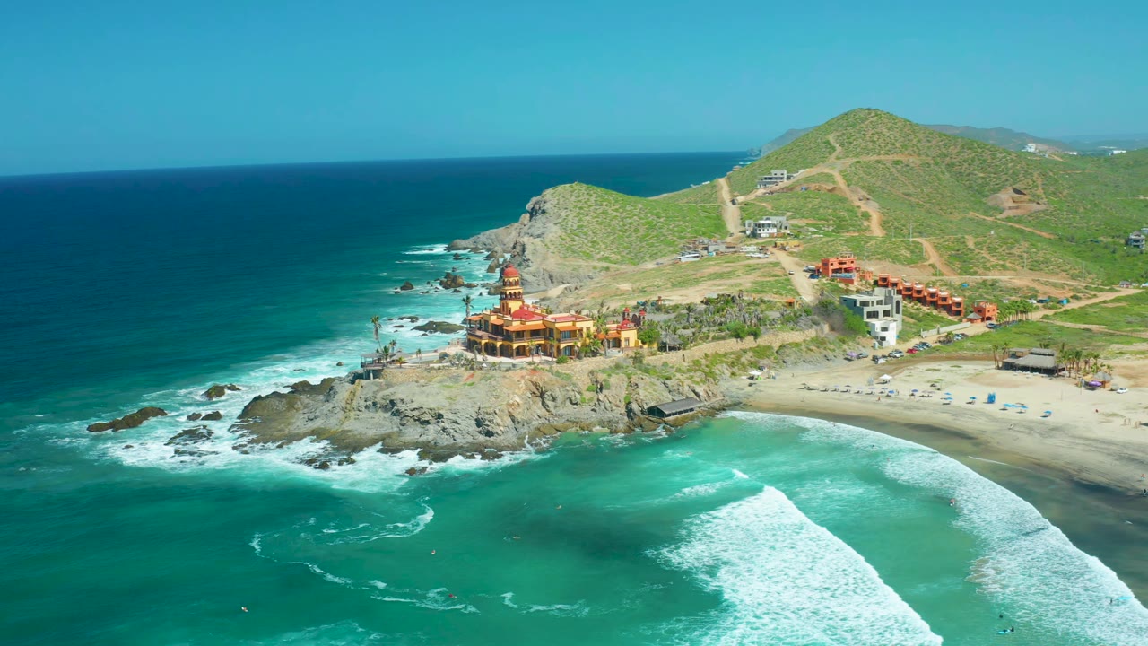 High aerial view of waves breaking around rocks beside a sandy beach and hillside buildings.