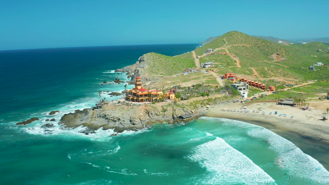 Aerial frame at Cerritos Beach showing the rocky point and temple-like building across the middle, a large surf line in the lower right water, green hills beyond, and no visible people.