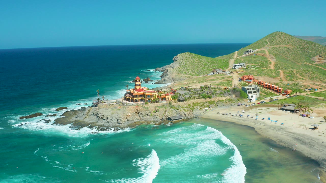 Aerial frame of Cerritos Beach in Baja California Sur with turquoise water, white foam, a rocky point, sandy beach, coastal buildings, green hills, and no visible people.