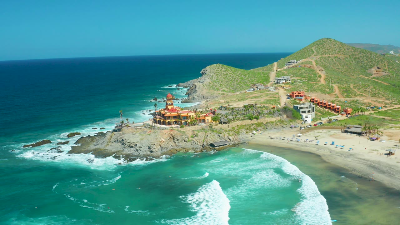 Aerial surf wraps around rocks below coastal buildings and green hills.
