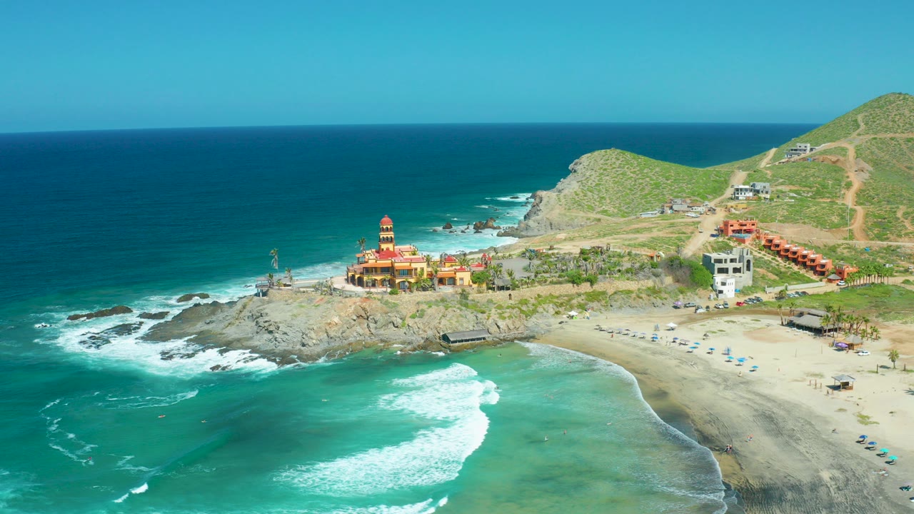 Opening aerial frame over Cerritos Beach, Baja California Sur, with turquoise surf in front, a rocky point and lighthouse-like building near center, sandy beach to the right, green hills behind, and no visible people.