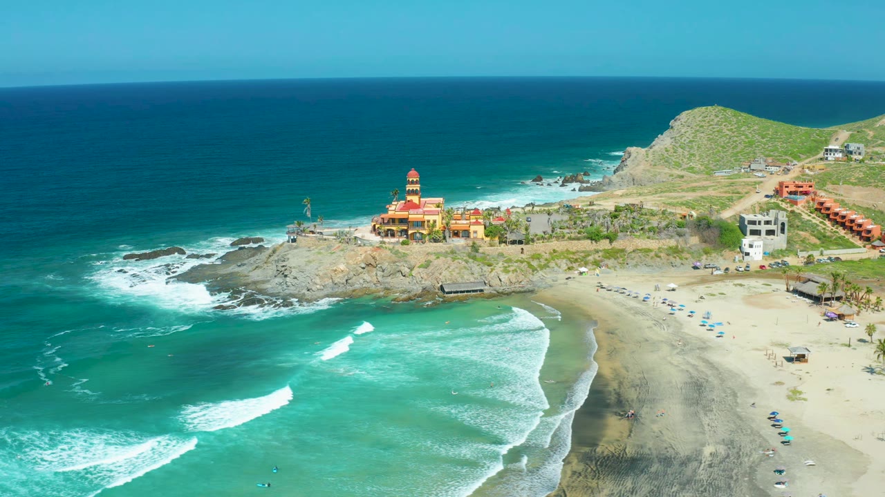 Aerial view at 0:01 over Cerritos Beach, Baja California Sur, with a broad band of ocean on the left, surf approaching the sand, a rocky point with a red-and-yellow towered building, hillside buildings, and no visible people.