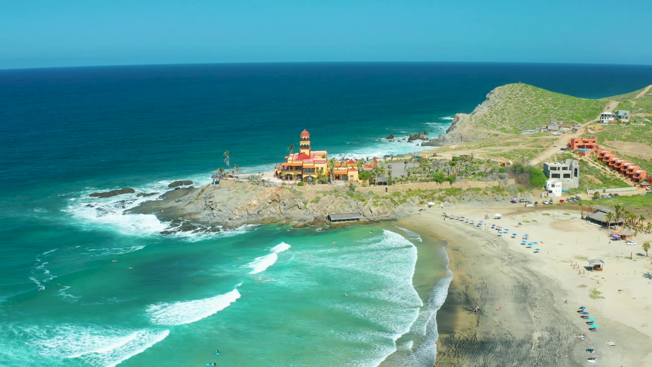 At 0:02, Cerritos Beach appears from above with white wave lines crossing turquoise water, a curved sandy beach, rocky headland, low green hills, scattered coastal buildings, and no visible people.