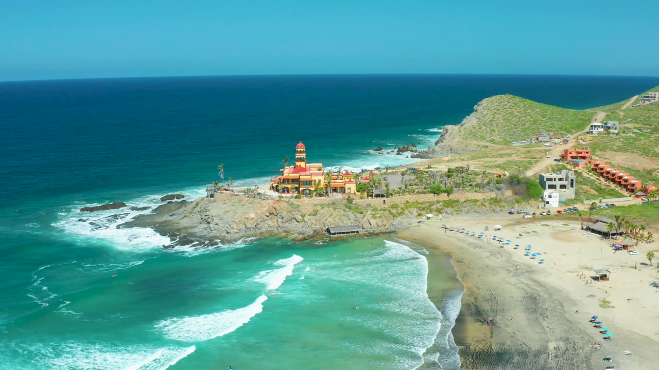 Aerial coastline view with sandy beach, waves, rocks, buildings, and green hills.