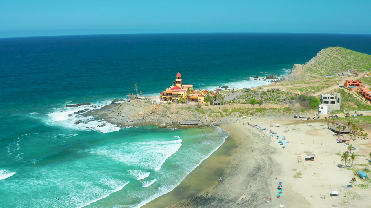 Opening aerial frame over Cerritos Beach, Baja California Sur, showing turquoise surf, pale sand, rocky point, cliffside buildings, a temple-like structure, a grassy hill, and no visible people.