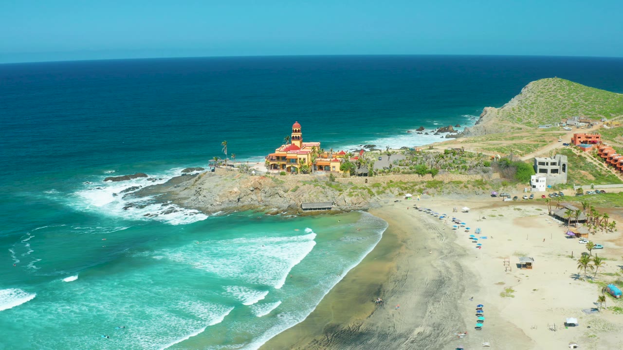 Cerritos Beach frame at 0:02 shows a broad turquoise wave band in the foreground, scattered blue umbrellas on the beach, cliffside buildings, a temple-like structure, and no visible people.
