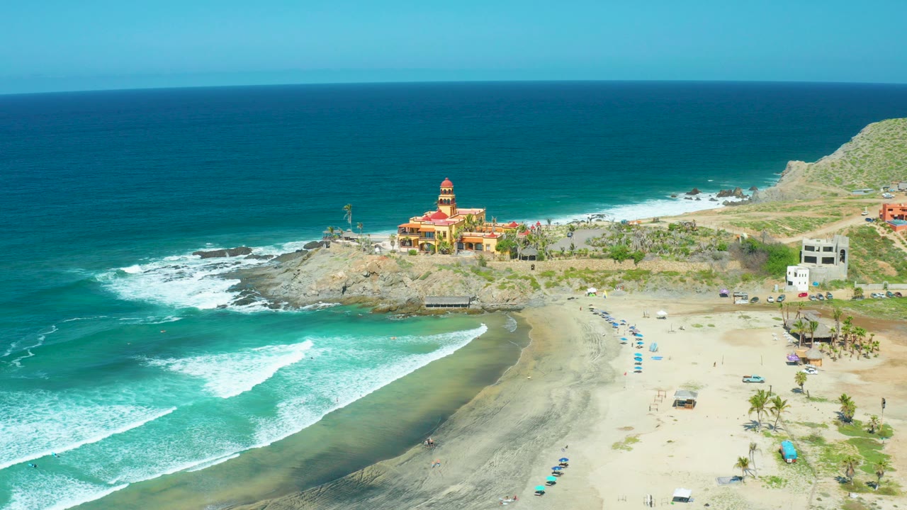 Aerial view at Cerritos Beach, Baja California Sur, with turquoise surf entering from the left, a sandy beach below, a rocky point with a yellow and red coastal building, green hillside, clear sky, and no visible people.