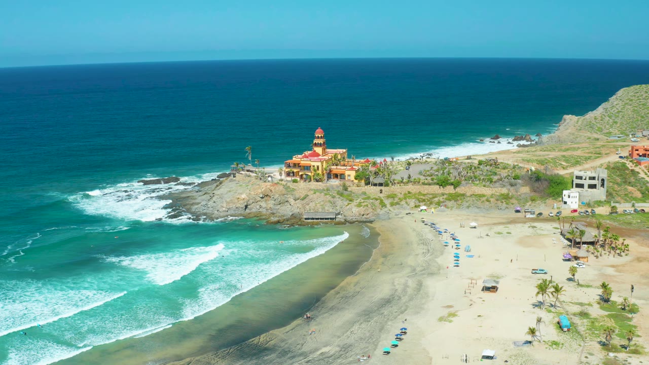 Aerial frame at Cerritos Beach showing white waves curling along the left edge, dark wet sand near shore, the yellow and red coastal building on the rocks, scattered umbrellas on the beach, and no visible people.