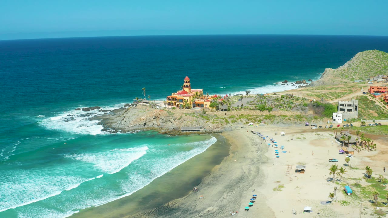 Aerial beach view with incoming surf and a rocky point with a coastal building.