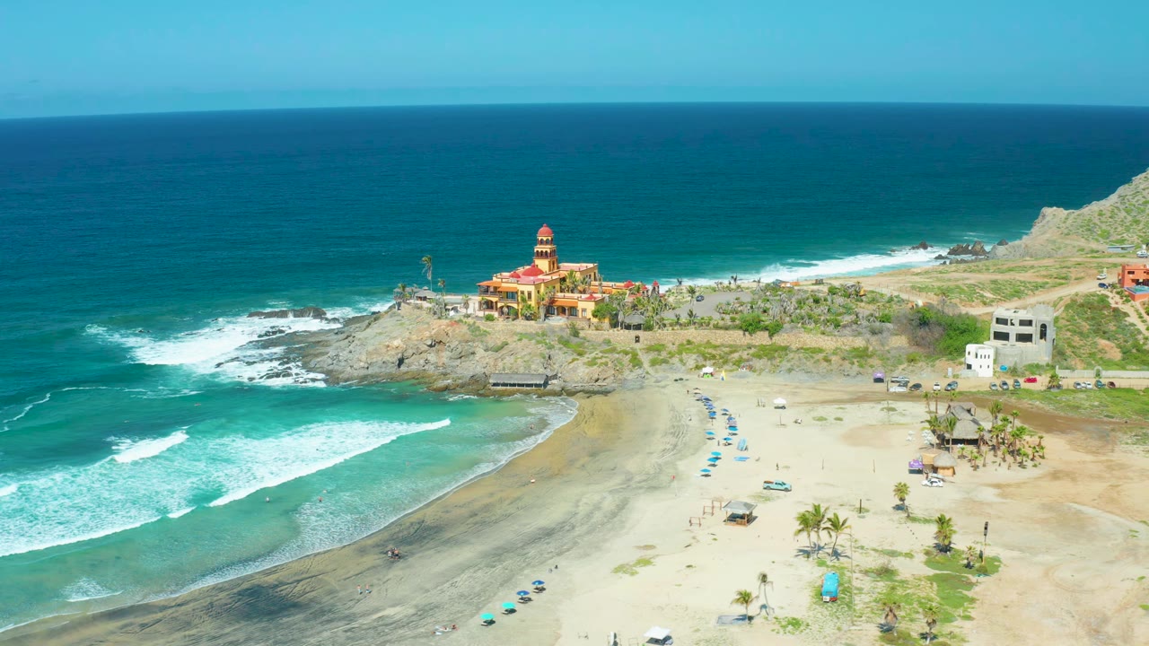 Cerritos Beach aerial frame with white waves breaking closer to the left shoreline, dark wet sand curving across the beach, blue umbrellas in rows, palms, rocky cliffs, and no visible people.