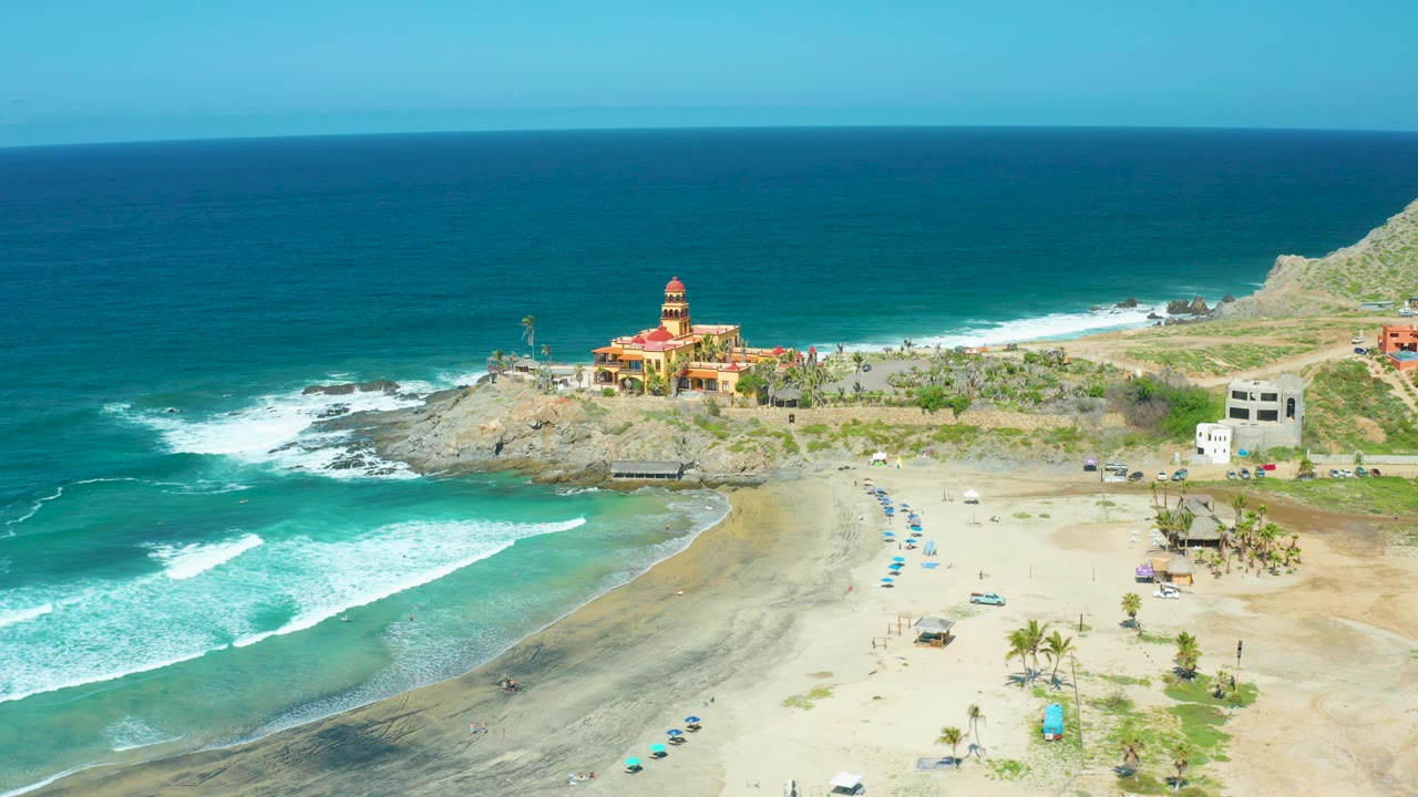 Aerial view over Cerritos Beach with a broader white surf band entering from the left, pale sand and beach umbrellas below, palms near the lower right, rocky cliffs, and no visible people.