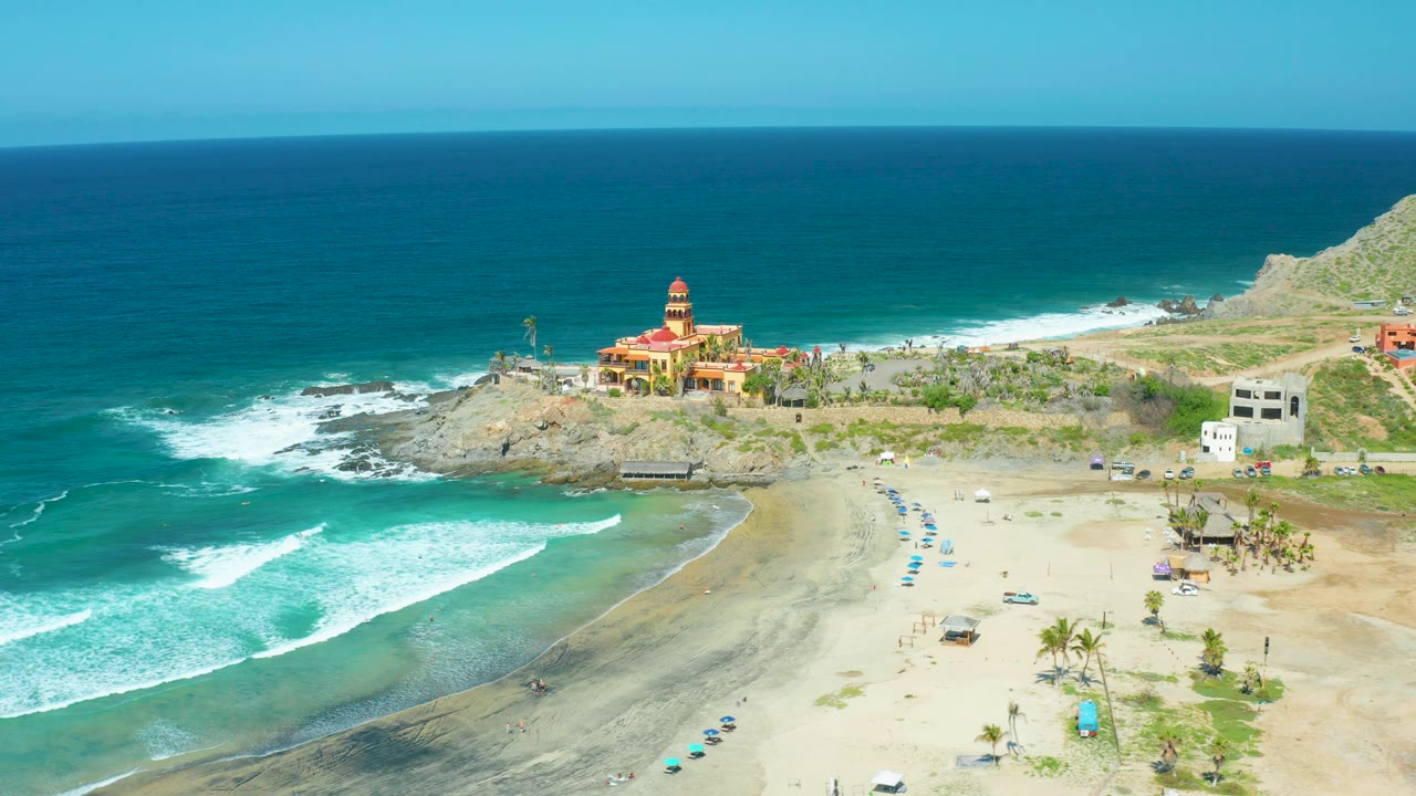 Cerritos Beach wide aerial frame with foamy waves spread along the left edge, dark shoreline sand in the lower center, beach umbrellas and palms to the right, cliffs and a red-roofed building beyond, with no visible people.