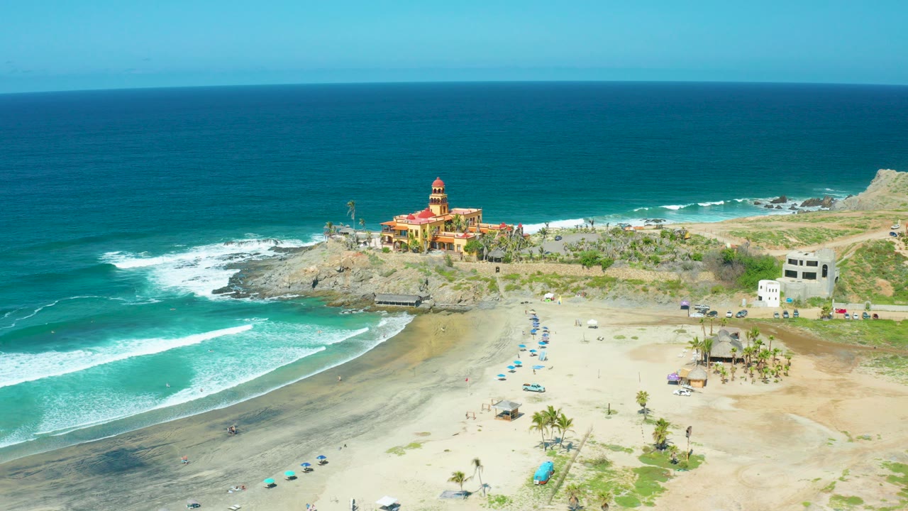 Aerial frame of Cerritos Beach with teal surf curling onto sand, blue umbrellas below, and a red building on the rocky point under a clear sky, with no recognizable people visible.