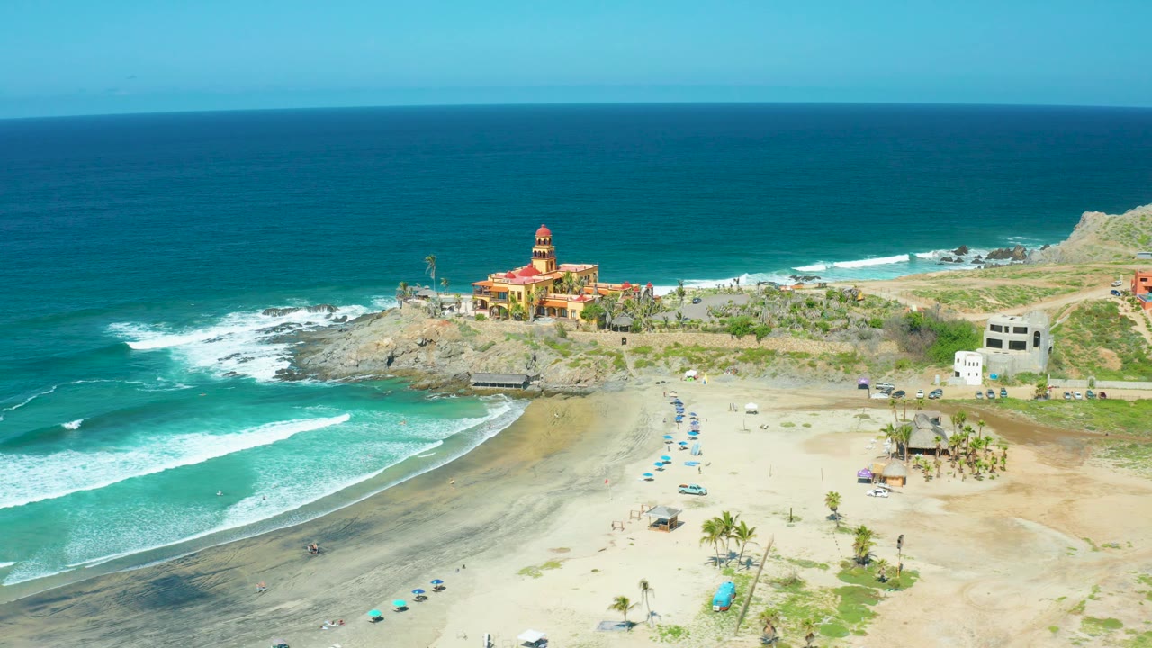 Aerial frame of teal water, beach umbrellas, and a red building on a rocky coast.