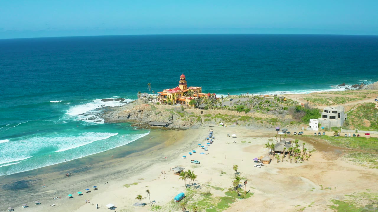 Aerial view of Cerritos Beach in Baja California Sur with turquoise water, white surf, sandy shore, umbrellas, rocky outcrops, coastal buildings, and no visible people.