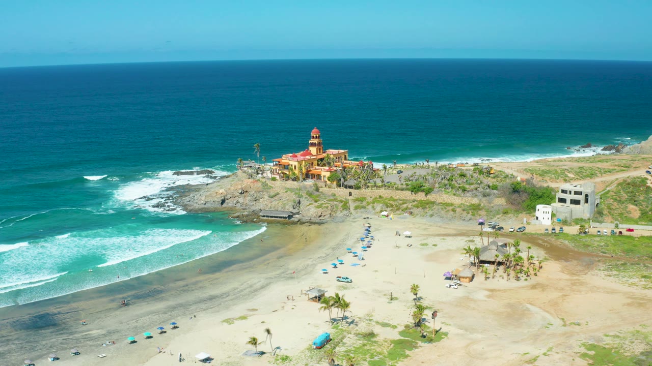 Wide Cerritos Beach view with the ocean horizon high in frame, white wave lines crossing the left foreground, sandy beach below, coastal buildings near the rocky point, and no visible people.