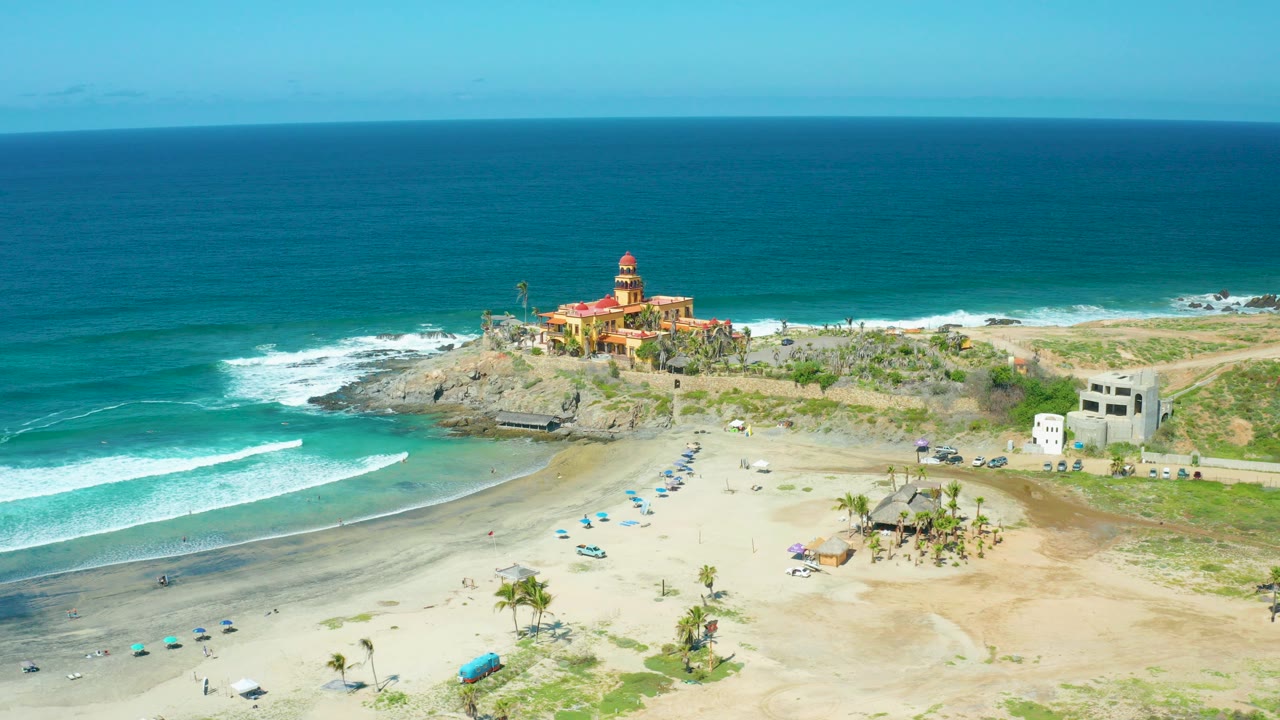 Aerial frame of Cerritos Beach, Baja California Sur, Mexico, showing a red-roofed lighthouse on a rocky point, turquoise ocean, breaking surf, umbrellas, sand, and tiny beachgoers.