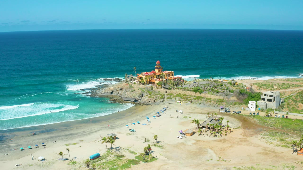 Early frame of Cerritos Beach in Baja California Sur from above, with white surf bands crossing turquoise water, scattered umbrellas on sand, small visible beachgoers, and a red-roofed building on the rocky point.