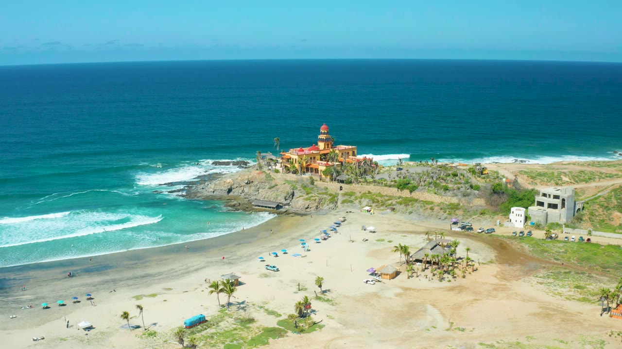Wide aerial view of Cerritos Beach surf rolling toward umbrellas and a rocky outcrop.