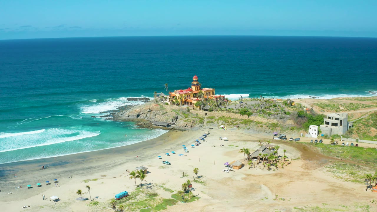 Cerritos Beach in a high wide frame, showing open sand in the lower half, white wave foam to the left, small visible people near the umbrellas, and the coastal building and rocks below a clear horizon.