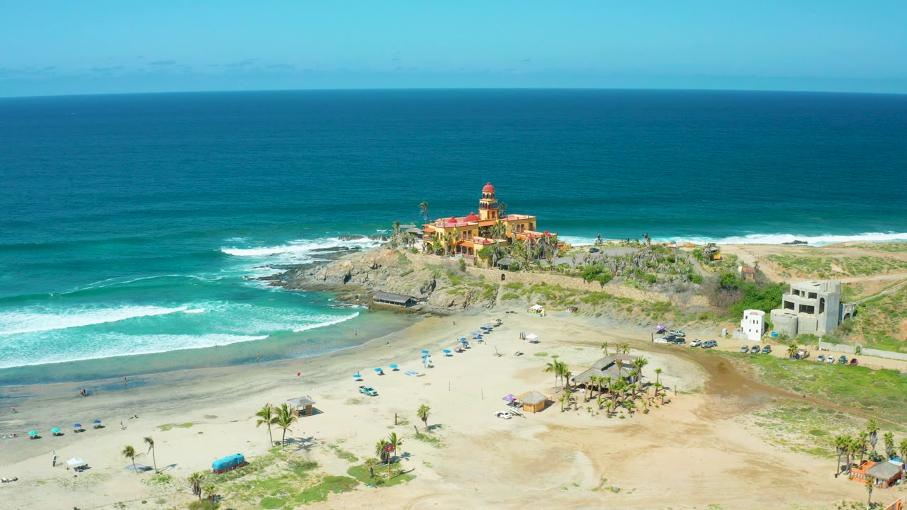 Opening aerial view at Cerritos Beach, Baja California Sur, with a red-topped lighthouse on a rocky point, surf curling toward the sand, blue umbrellas, and no visible people.