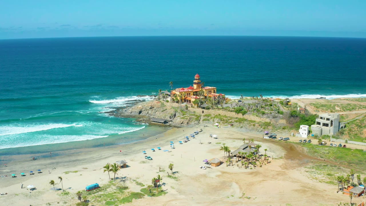 Lighthouse above rocky coastline with surf, sandy beach, umbrellas, and palms.