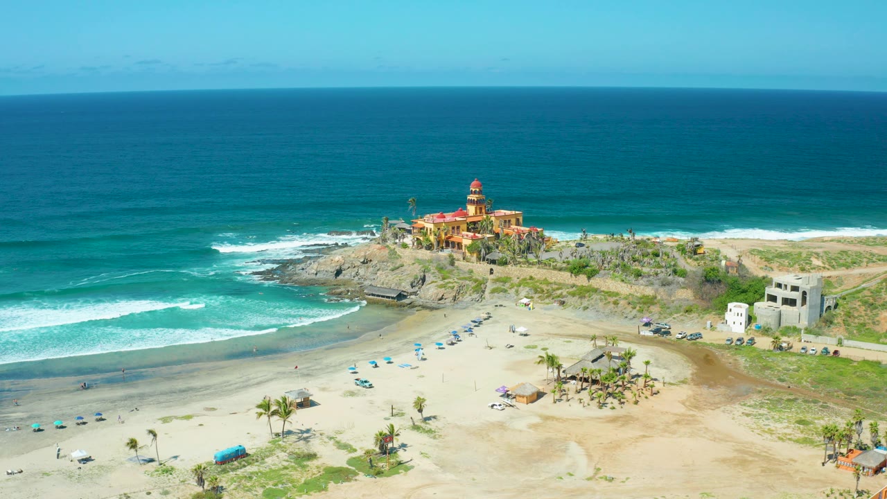 A wide aerial frame at Cerritos Beach with the lighthouse on the rocky headland, turquoise water darkening toward the horizon, pale sand in front, and no visible people.