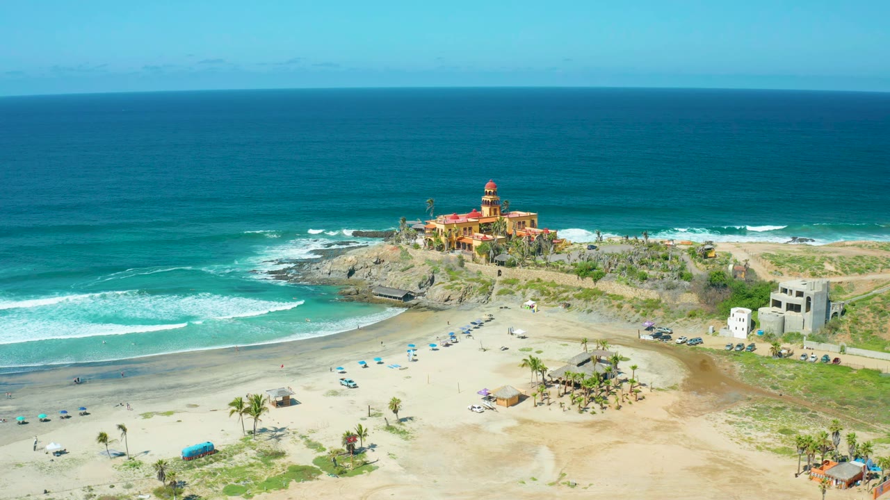 Cerritos Beach wide aerial view showing the lighthouse slightly right of center, surf wrapping the rocky point, blue umbrellas on sand, palms, small buildings, and no visible people.