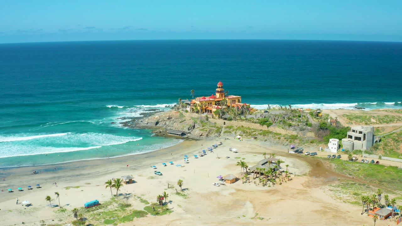Cerritos Beach aerial view with the lighthouse on the rocky headland, bands of white surf at left, open sand and blue umbrellas in the foreground, small buildings at right, and no visible people.