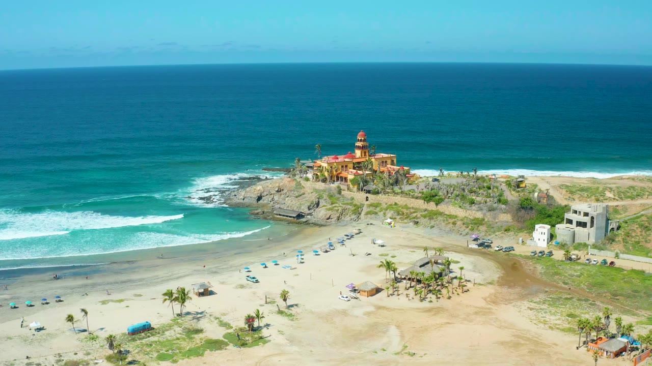 At Cerritos Beach in Baja California Sur, Mexico, a wide aerial frame shows turquoise ocean, white surf, tan sand, umbrellas, parked cars, palm trees, buildings, and a lighthouse on a rocky outcrop, with no visible people.