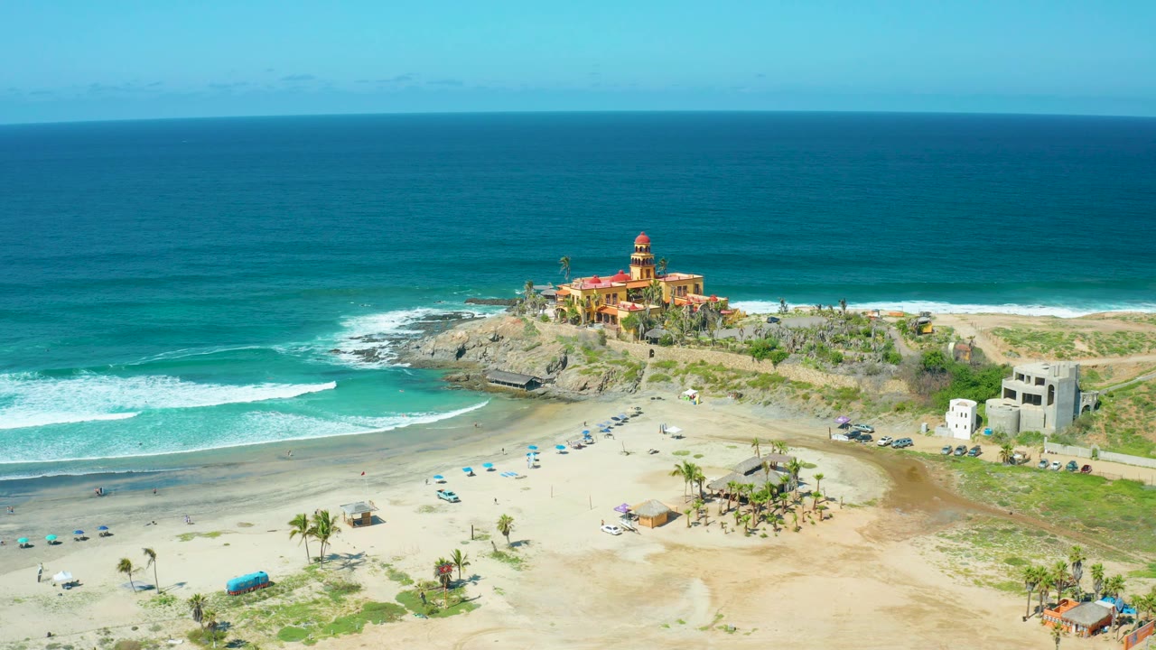 At Cerritos Beach, Baja California Sur, Mexico, bright sunlight shows white foam sweeping across the left side of the beach while cars, umbrellas, palms, low buildings, and the lighthouse point remain visible, with no visible people.