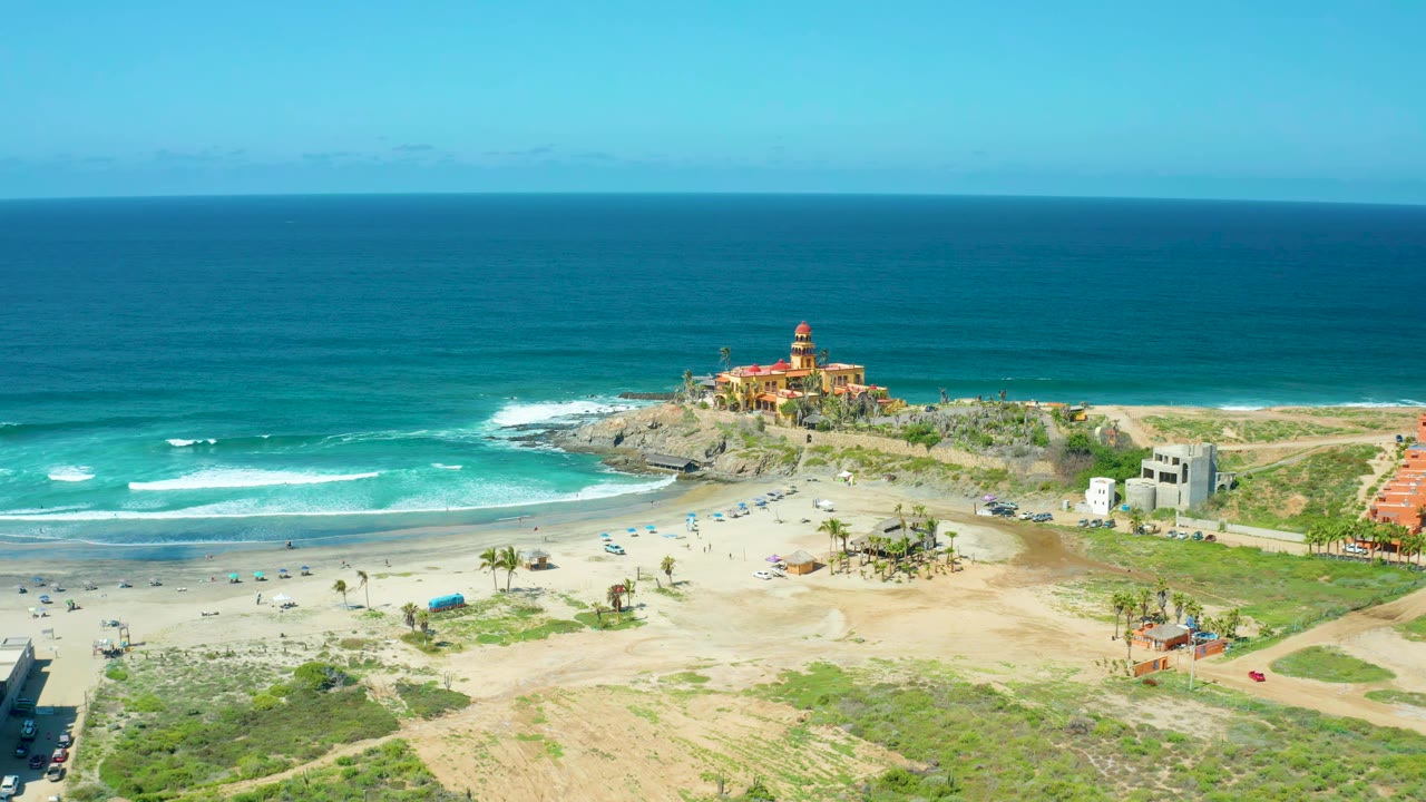 Wide aerial frame of Cerritos Beach with the horizon high in the image, waves curling toward the sand, beach umbrellas below, and the lighthouse-like structure centered on the rocky point, with no visible people discernible.