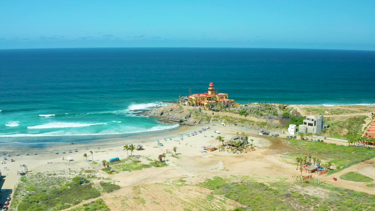 Aerial view over Cerritos Beach showing open sand in the lower frame, palms and umbrellas near the beach, blue-green water offshore, and the lighthouse-like building on the rocky headland, with no visible people discernible.