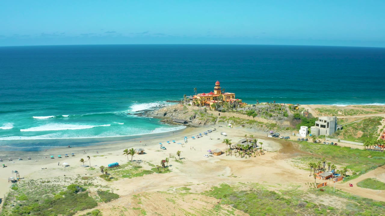 Elevated panorama of Cerritos Beach with darker blue ocean beyond the surf, pale sand and rough ground in the foreground, a cluster of palms and umbrellas near the center, and the rocky point building to the left, with no visible people discernible.
