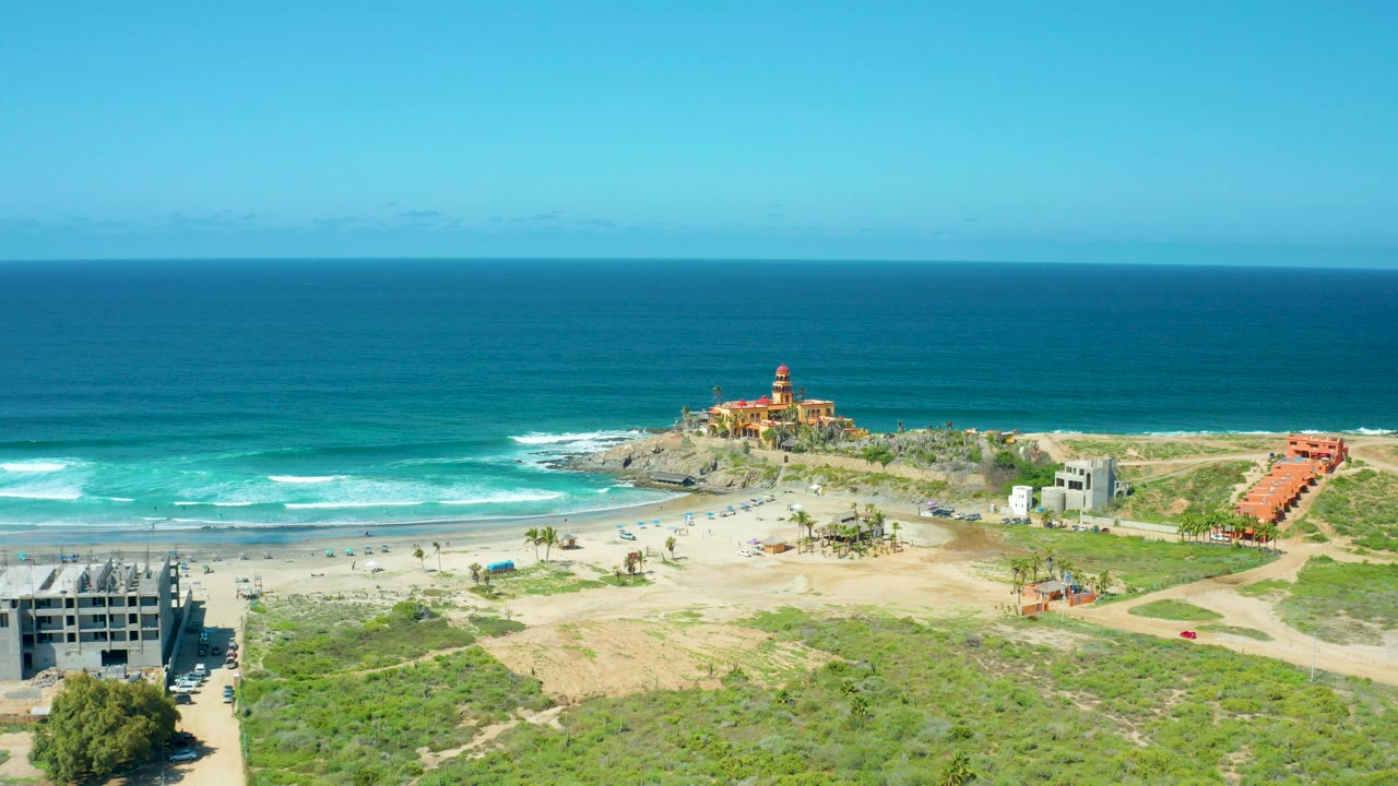 Aerial frame of Cerritos Beach, Baja California Sur, with turquoise ocean, white surf, sandy beach, a lighthouse-like building on a rocky point, small beachgoers, vehicles, dunes, and construction at the edges.