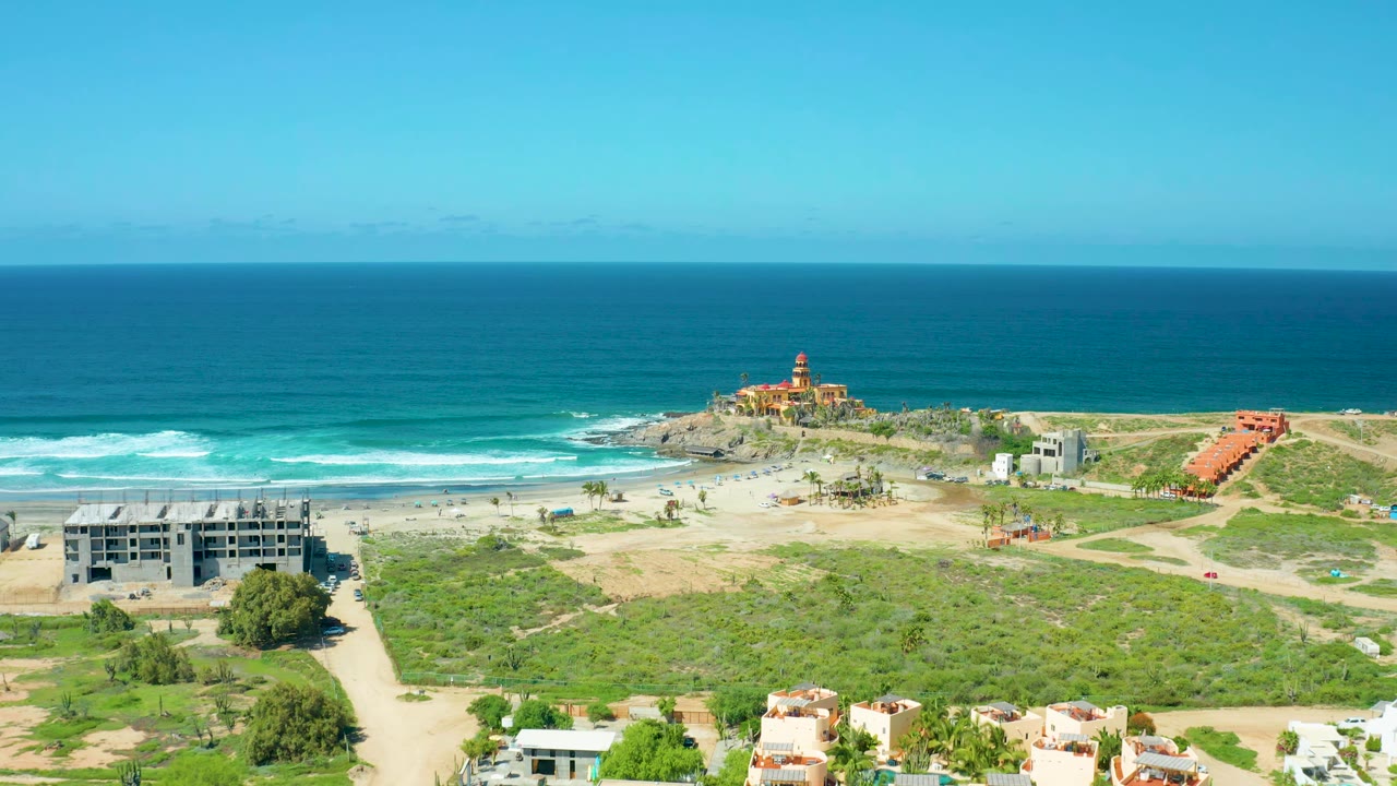 Wide aerial coastline view with breaking waves, sandy shore, green scrub, buildings, and a lighthouse-like structure.