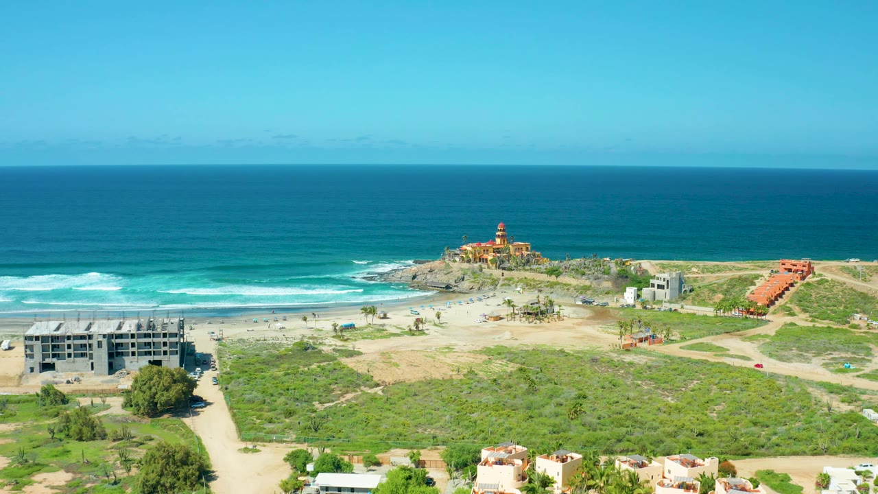 At Cerritos Beach, a broad band of blue ocean and whitecaps sits behind the sandy shoreline, with green coastal vegetation, a construction-like building at left, orange-roofed structures at right, and no visible people.