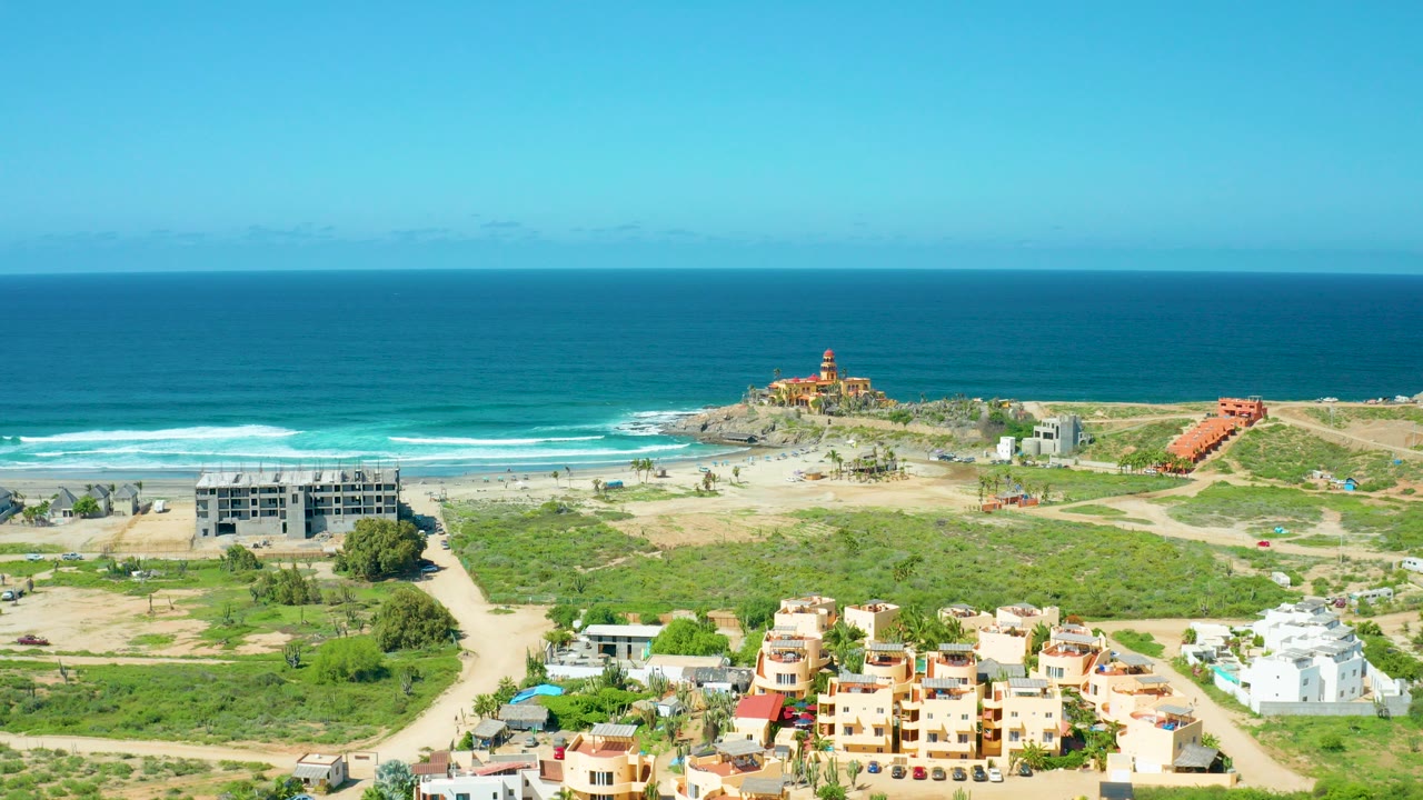 Opening aerial frame over Cerritos Beach, Baja California Sur with turquoise ocean, surf lines, low buildings, dirt roads, scrub greenery, and a lighthouse-like structure on the point, with no visible people.