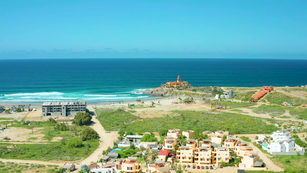 Wide drone view of coastal buildings, green lots, beach surf, and blue ocean.