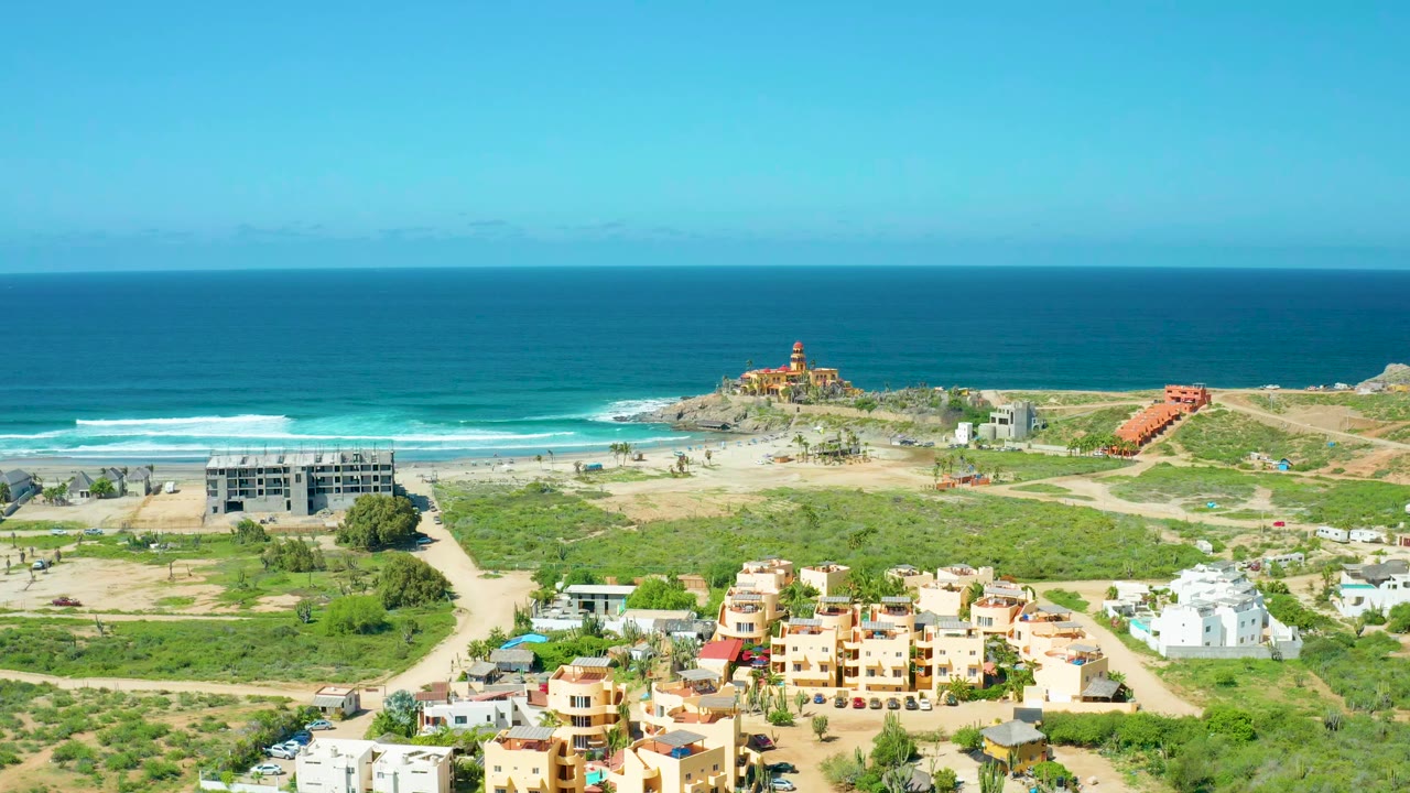 Coastal aerial view with low buildings, dirt roads, green scrub, beach, surf, and open ocean.