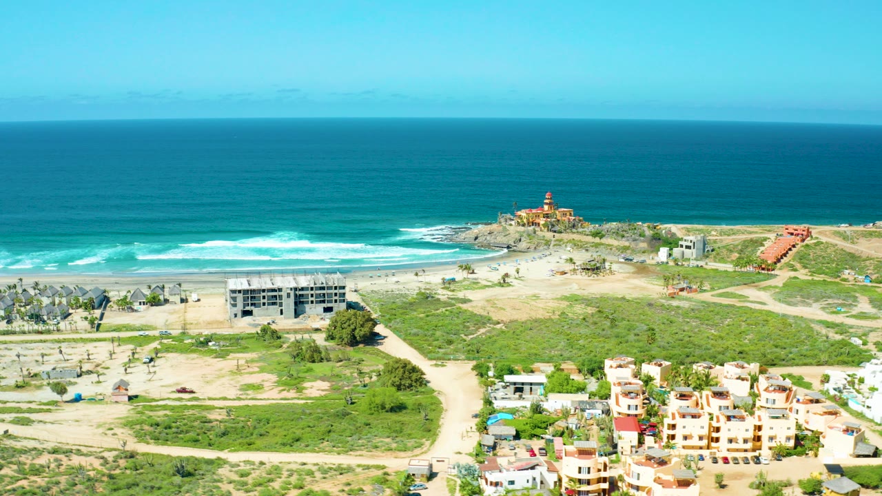 Cerritos Beach, Baja California Sur aerial view with turquoise ocean, white surf, sandy beach, low homes, green scrubland, and a lighthouse on a rocky outcrop, with no visible people.