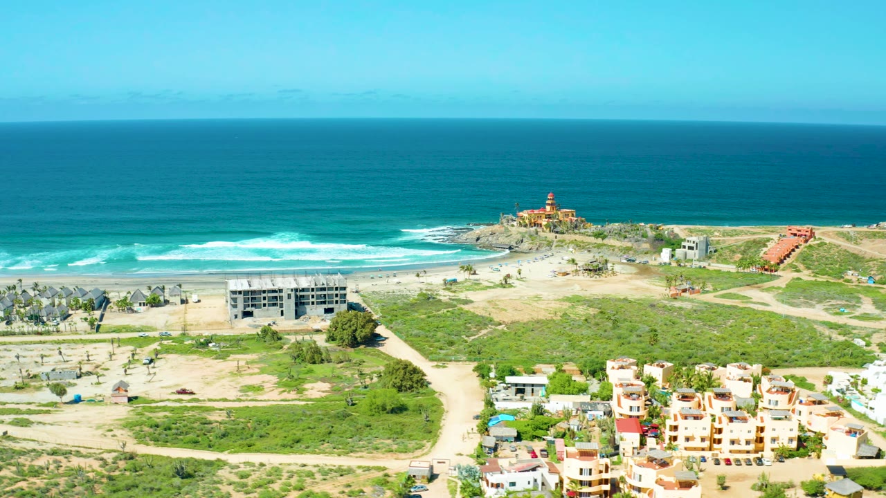 Wide coastal aerial with waves breaking near a lighthouse and homes in the foreground.