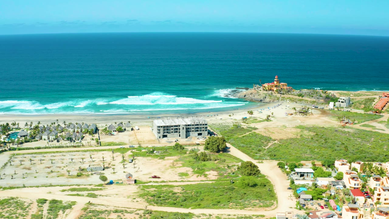 A high aerial view of Cerritos Beach, Baja California Sur shows a broad ocean horizon, breaking waves, the lighthouse point slightly right of center, sandy tracks, buildings, and no visible people.