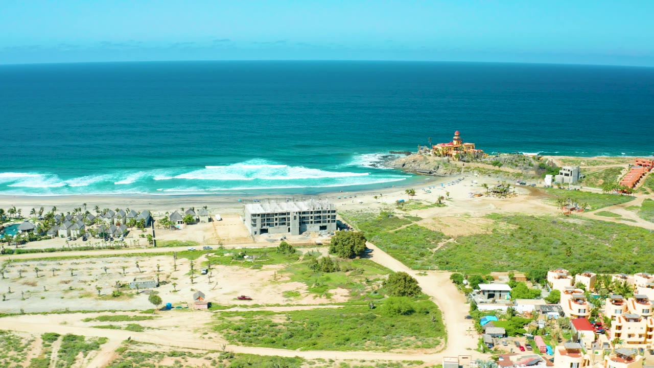 Cerritos Beach, Baja California Sur appears from above with the lighthouse near the right edge, foamy surf below the horizon, pale sand, green patches, residential buildings, and no visible people.