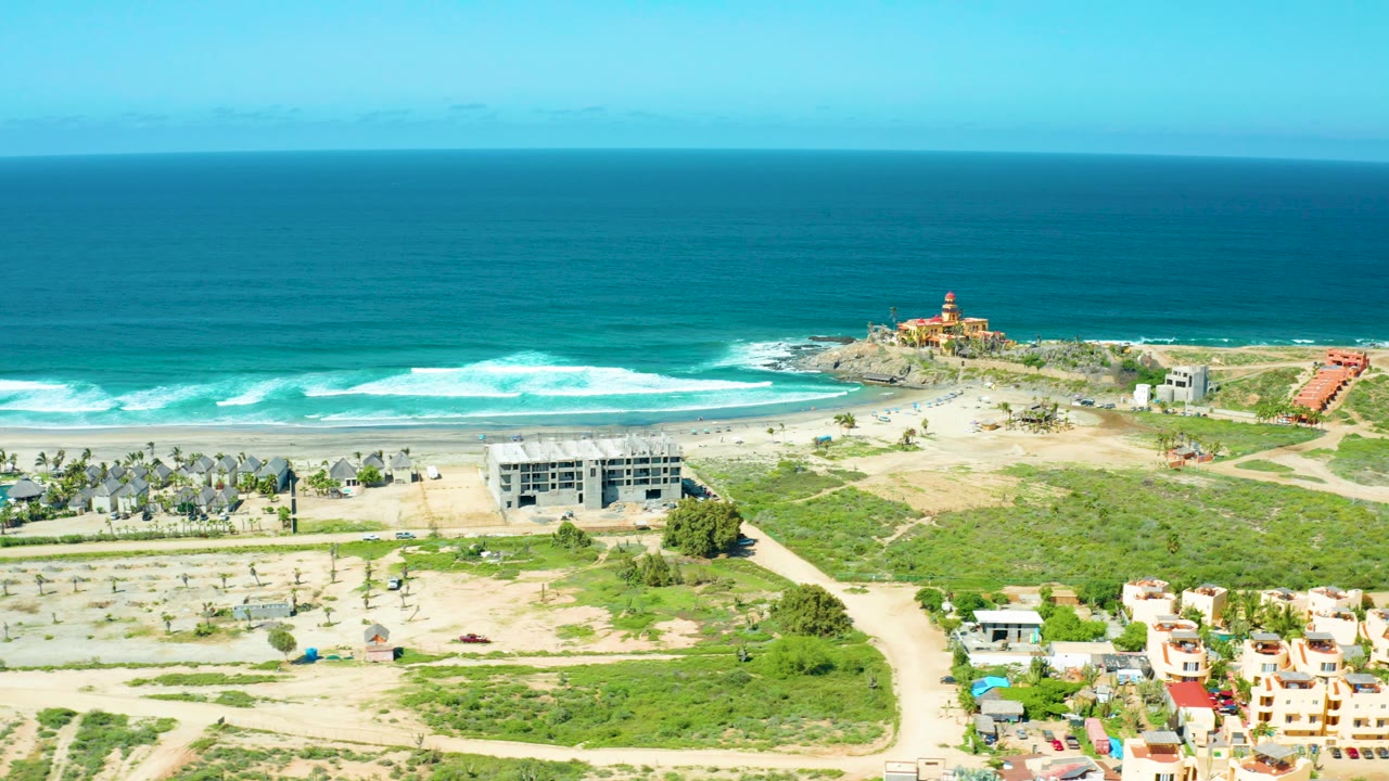 Drone view across beach lots, road, ocean waves, and a lighthouse on the coastal point.