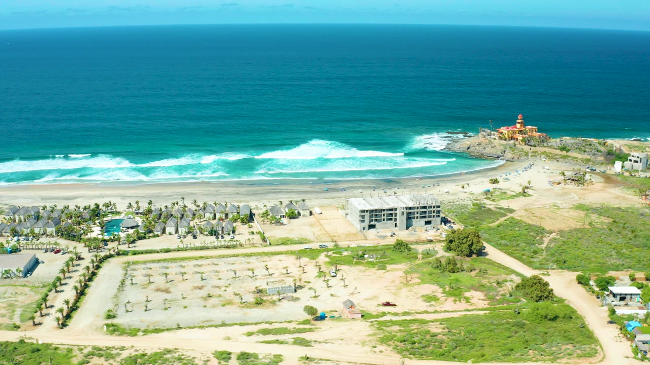 Wide aerial frame over Cerritos Beach, Baja California Sur, Mexico showing turquoise ocean, white surf, sandy shore, tents, dirt paths, greenery, low buildings, and no visible people.
